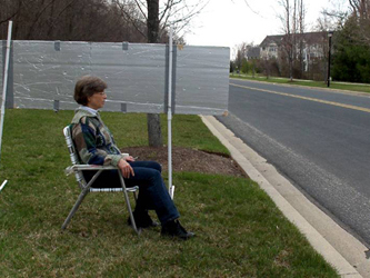 We are looking to the left along a straight two-lane street with a curb and grass on both sides, houses on the right side and woods on the left.  A woman is sitting in a lawnchair on the grass about 4 feet from the left curb, facing the street. A few feet beside her is a grey board suspended on two poles.  The board is 8 feet wide, the top is 5 feet above the ground, the bottom is about 3 feet above the ground, so that the board is beside the woman's head and shoulders.